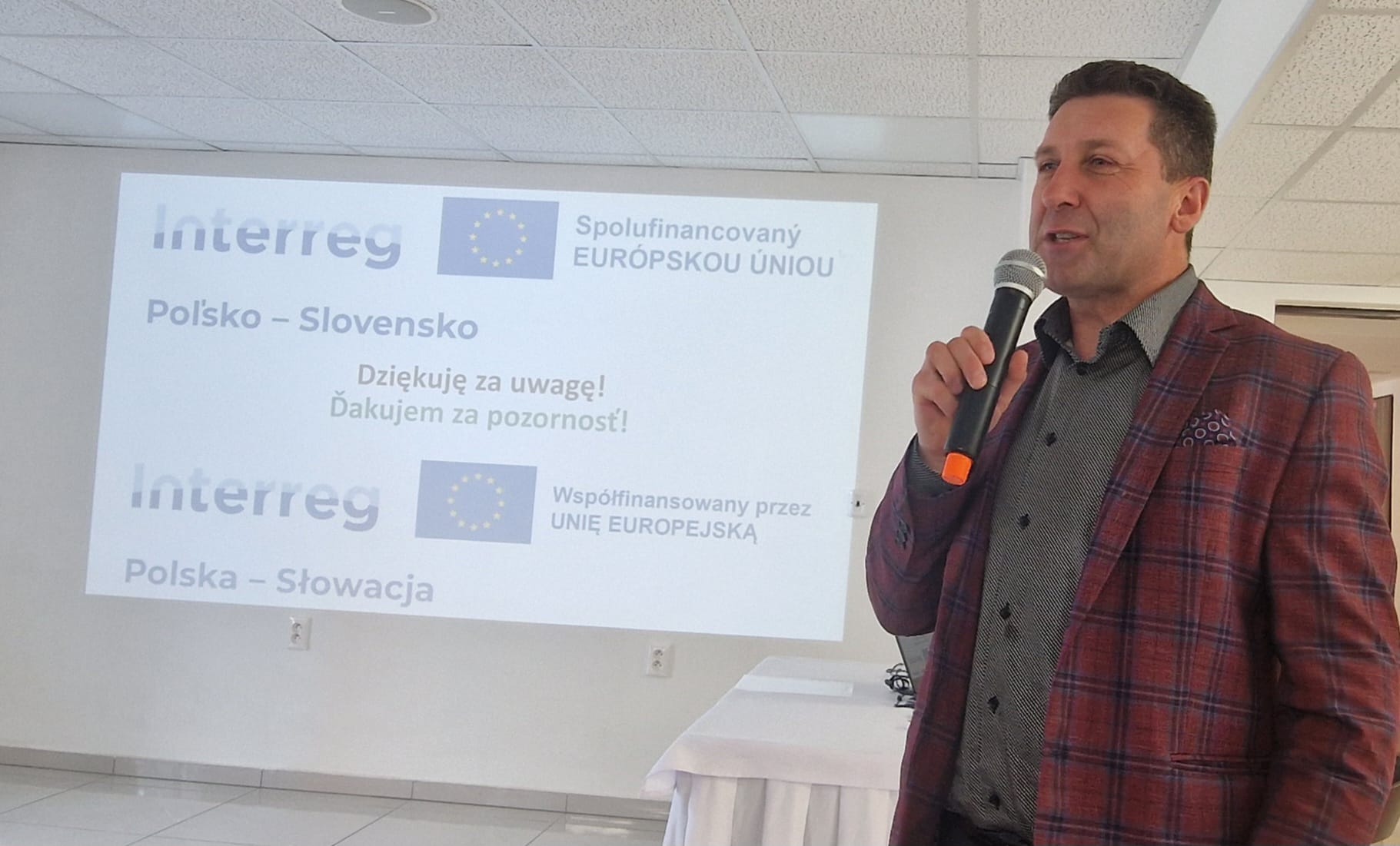 Mężczyzna z mikrofonem, w garniturze w kratę, przemawia na tle prezentacji Interreg Polska-Słowacja, finansowanej przez UE.